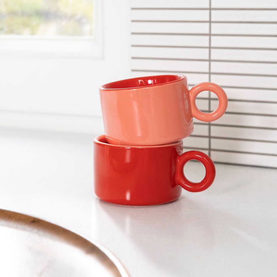 Two stacked ceramic mugs in red and pink on a white surface with a blurred background.