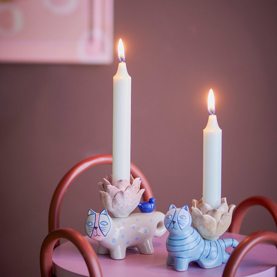Two lit candles in decorative holders on a small round table with a pink surface and brown legs against a purple wall.