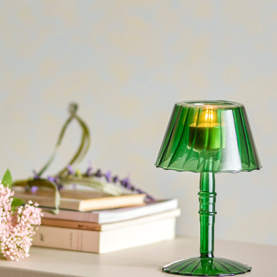 Green glass Votive holder on a white surface with books and flowers in the background
