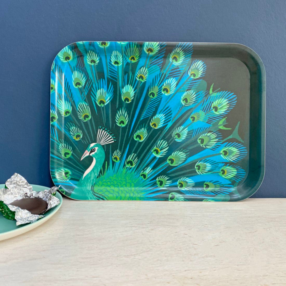 Peacock by Asta Barrington, peacock in full tail-feather on indigo background, 27x20 cm small rectangular tray, styled leaning against a blue painted wall, with a plate.