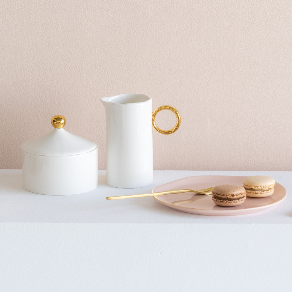 Good Morning white porcelain milk jug with wavy rim and gold ring handle, and white porcelain sugar bowl with gold sphere knob, styled with a Good Morning pink plate with two macarons and a gold fork.
